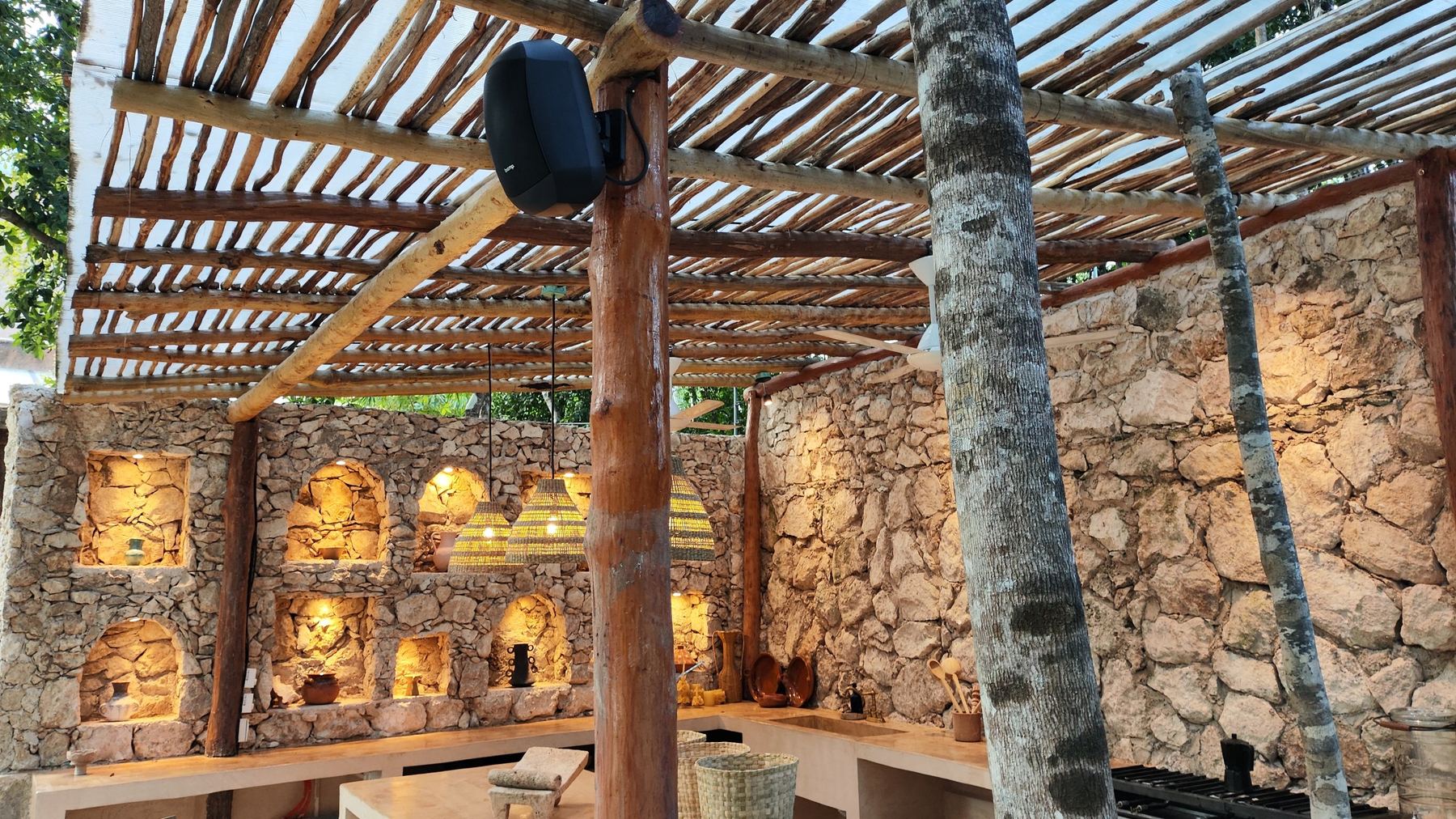 Bocina de exterior bajo una pérgola de madera, junto a muros de piedra iluminados